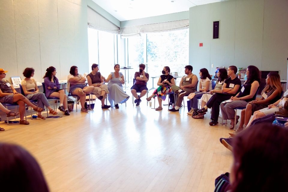 former interns sitting in a circle in front of a sun-filled window listening to a person speaking